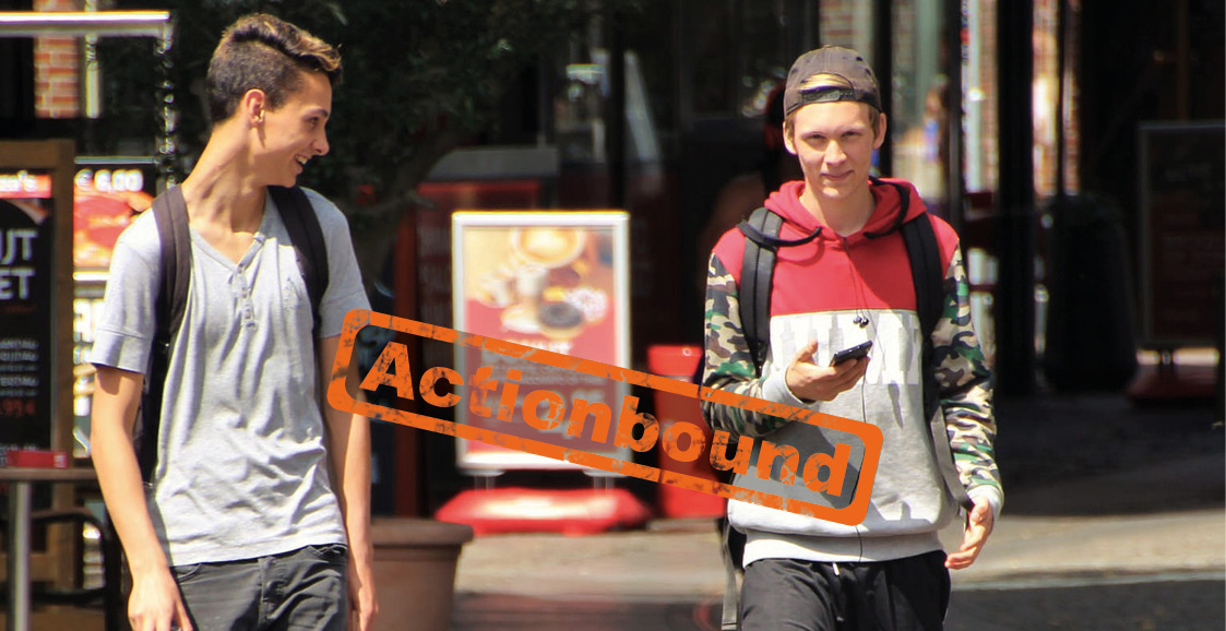 Zwei Jungen mit Handy sind in einer Stadt unterwegs, Stempel Actionbound auf dem Bild
