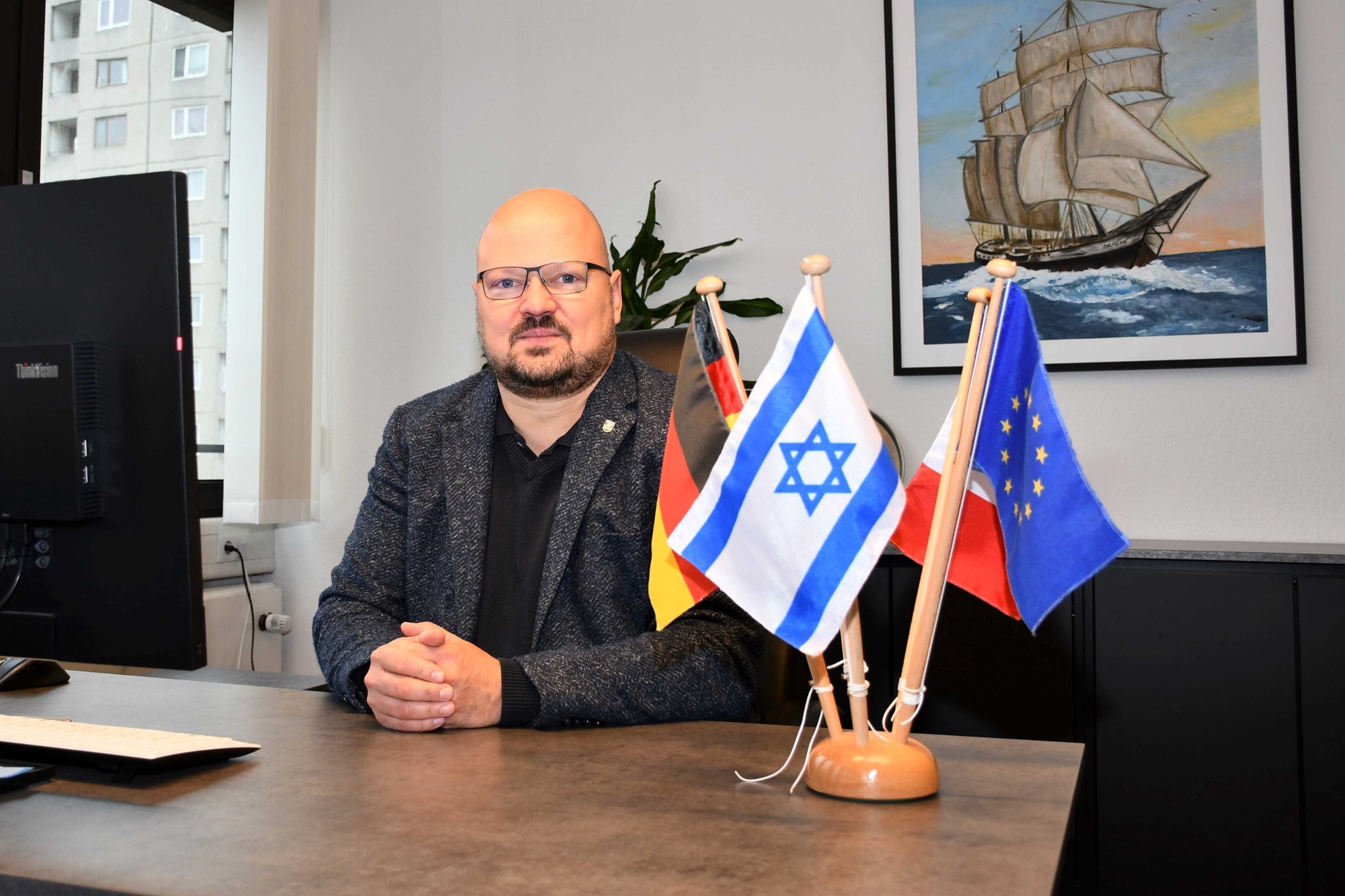 Bürgermeister Kai Eggert sitzt ernst an seinem Schreibtisch, auf dem ein Tischflaggenset mit der israelischen Flagge steht.