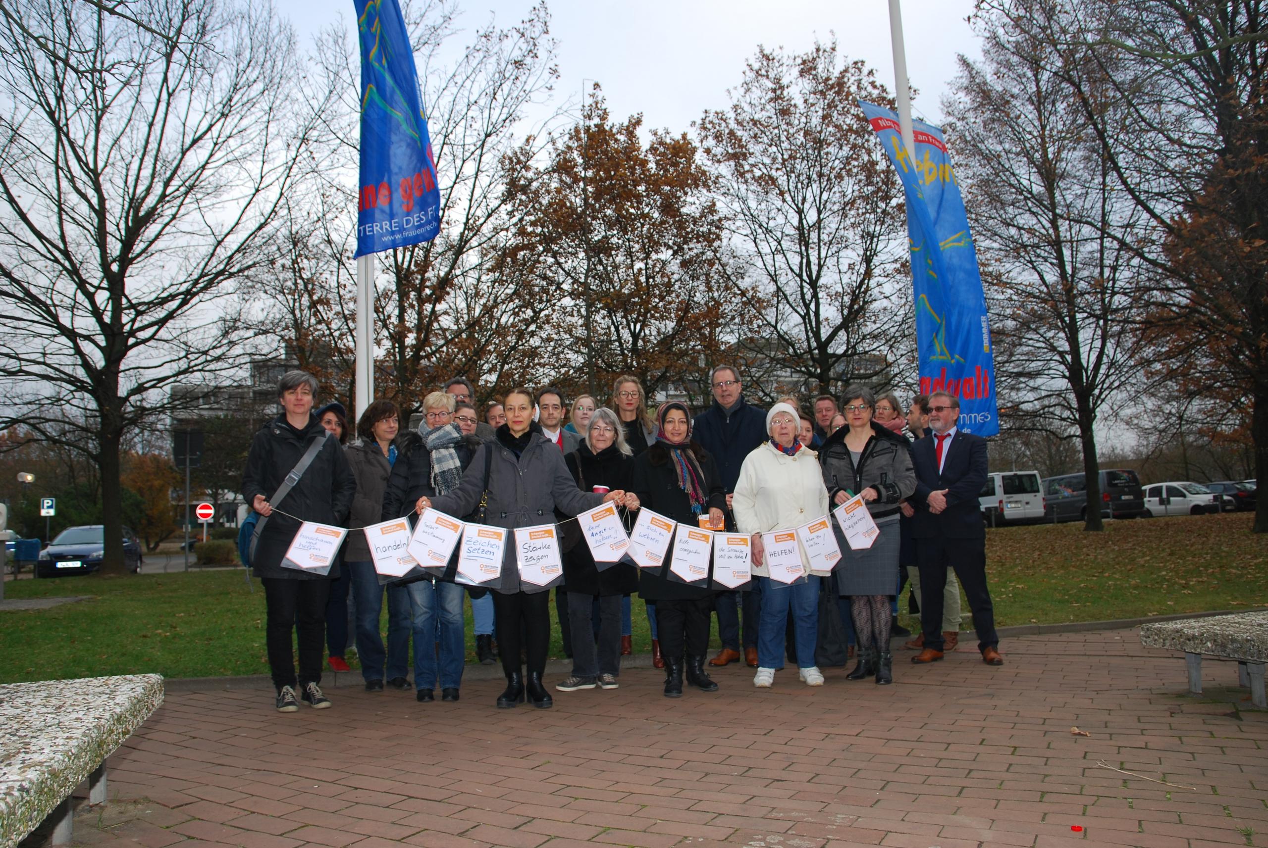 Menschengruppe vor gehissten Fahnen am Tag gegen Gewalt an Frauen