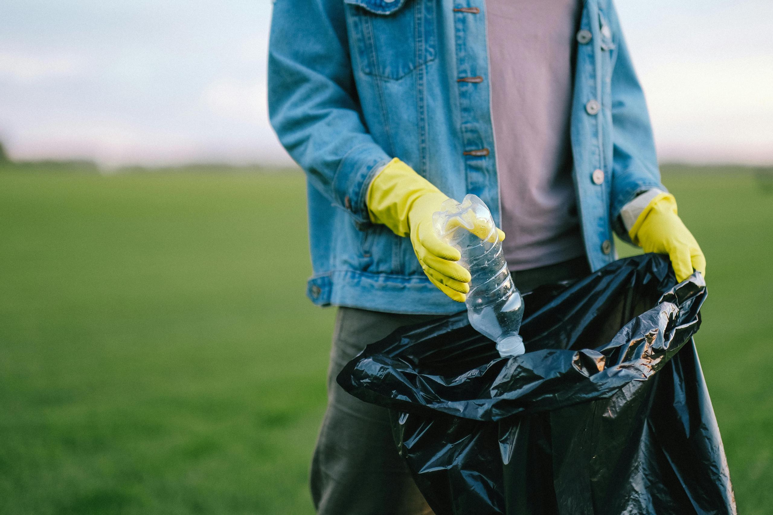 Ein Mann steht auf einer grünen Wiese. In der einen Hand hält er einen schwarzen Müllbeutel, in den er eine Plastikflasche steckt.