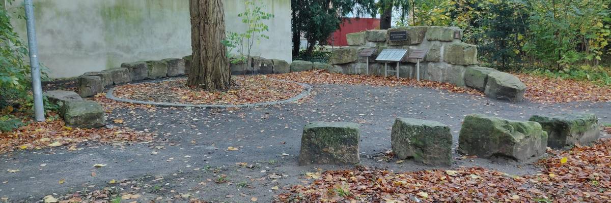 Kreis aus unbehauenen Natursteinen auf der Rückseite vom Ehrenmal in Alt-Laatzen. An einem Punkt des Kreise sind mehrere der unbehauenen Natursteine aufeinander gestapelt. An den Steinen ist zentral ein Bronzeschild angebracht. Davor steht zentral eine Metallkassette mit Toten Gedenksteine. Rechts und links von der Metallkassette steht je eine Bronzetafeln mit Inschrift.