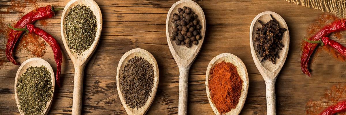 Holzkochlöffel, die mit unterschiedlichen Gewürzen gefüllt sind liegen geordnet auf einem Holztisch. Chilischoten liegen darum herum.