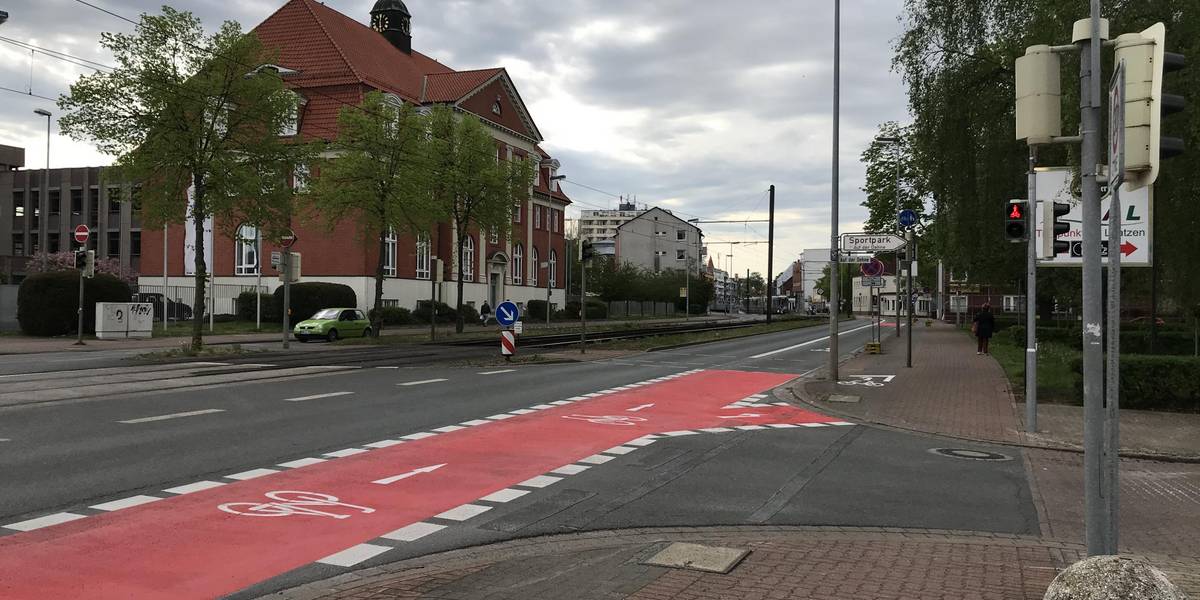 Roter Fahrradstreifen auf einer Straße. Rechts davon sind Geschäfte zu sehen.