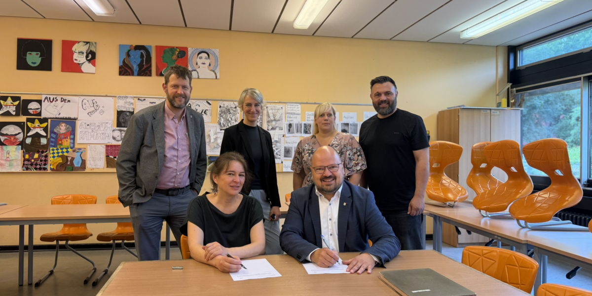Bürgermeister Kai Eggert und Planerin Kristina Gronau unterzeichnen in einem Klassenraum den Vertrag zur Sanierung des C-Trakts der Albert-Einstein-Schule. Hinter ihnen stehen weitere Vertreterinnen und Vertreter von Schule und Stadtverwaltung.