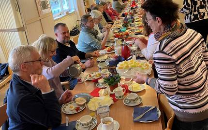 Eine Gruppe von Menschen sitzt an einem langen Tisch und frühstückt. Der Tisch ist gedeckt mit Obst, Brötchen und weiteren Lebensmitteln.