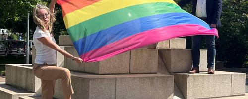 Ein Mann und eine Frau stehen auf Steinstufen. Zwischen ihnen weht eine Regenbogenflagge, die beide in ihren Händen halten.