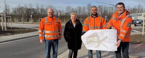Drei Männer in orangenen Warnjacken und eine Frau stehen an der Einmündung einer neuen Straße auf dem Gehweg.