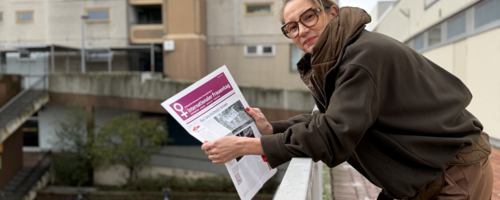 Eine Frau mit festem Blick lehnt über eine Brüstung, in der Hand hält sie eine Zeitung mit violettem Deckblatt.