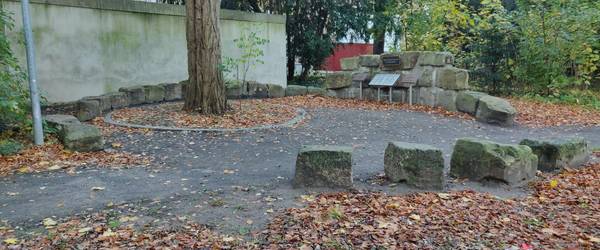 Kreis aus unbehauenen Natursteinen auf der Rückseite vom Ehrenmal in Alt-Laatzen. An einem Punkt des Kreise sind mehrere der unbehauenen Natursteine aufeinander gestapelt. An den Steinen ist zentral ein Bronzeschild angebracht. Davor steht zentral eine Metallkassette mit Toten Gedenksteine. Rechts und links von der Metallkassette steht je eine Bronzetafeln mit Inschrift.