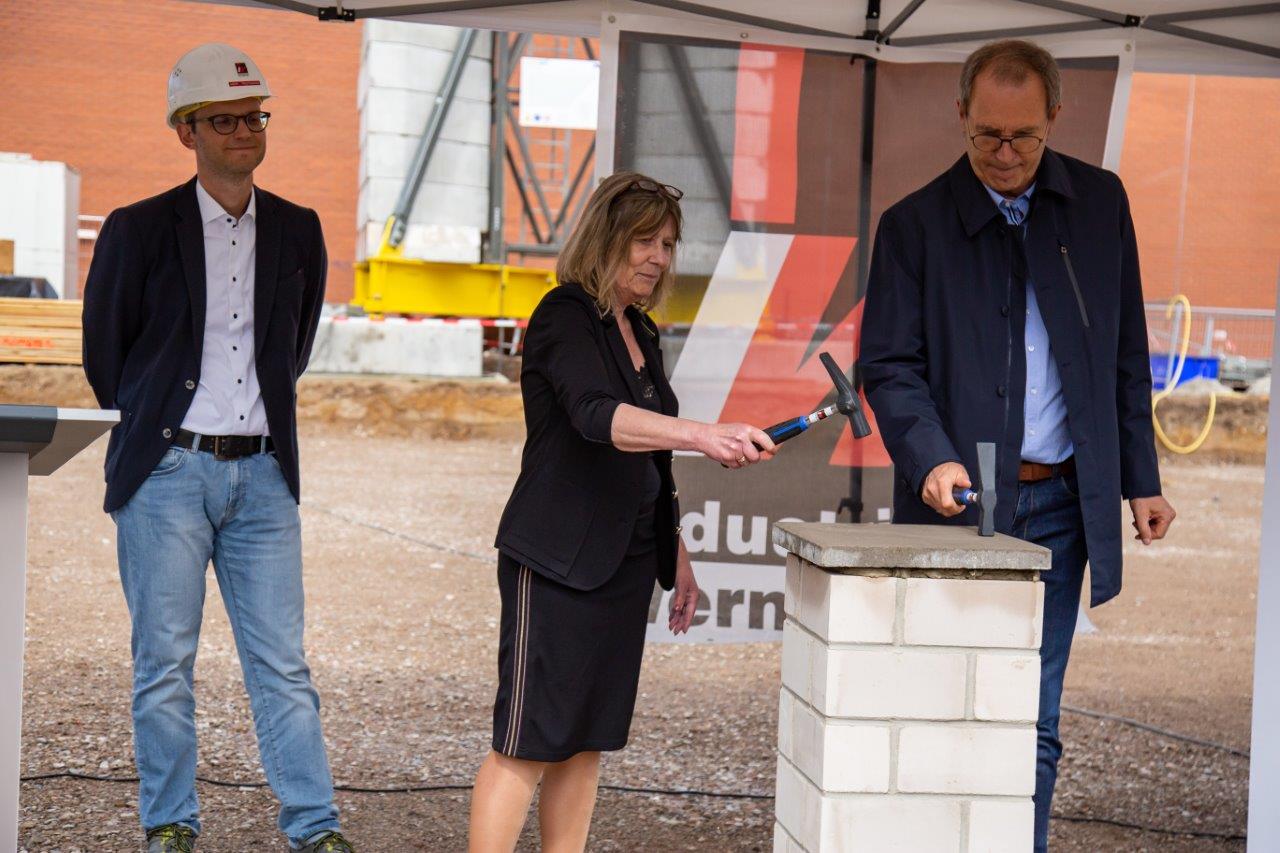 Im Vordergrund stehen Bürgermeister Köhne und Schulleiterin Kohl, die Grundmauer verschließen. Im Hintergrund stehen weitere Beteiligte und beobachten die beiden.