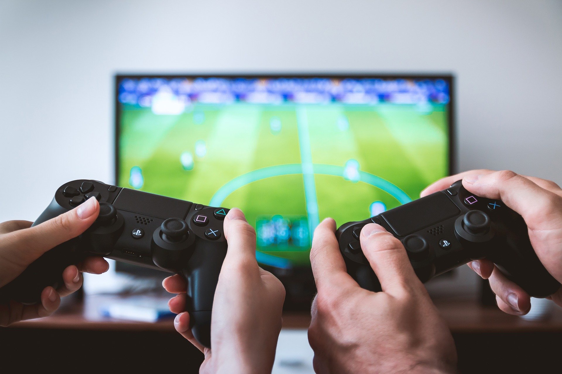 Es sind die Hände zweier Spieler zu sehen, die jeweils einen Controller halten und im Hintergrund befindet sich ein Fernseher mit FIFA
