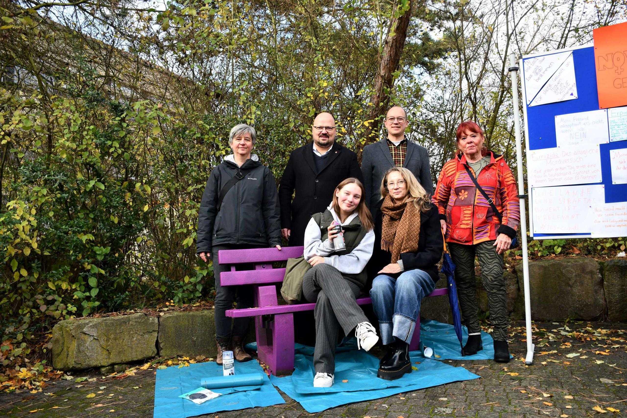 Zwei Männer und vier Frauen sitzen und stehen um eine lila Bank herum.