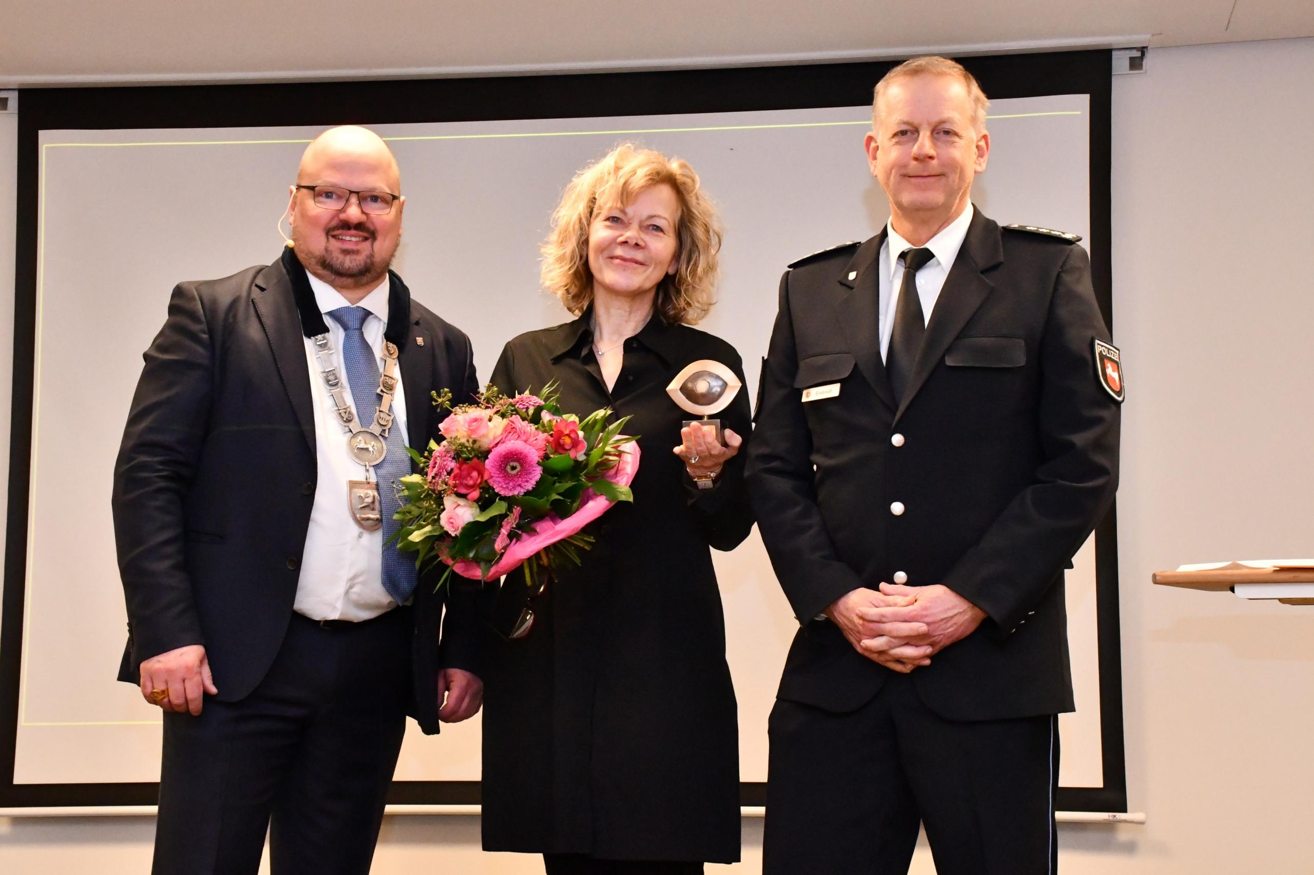 Laatzens Bürgermeister Kai Eggert, Corinna Luedtke und Leiter des Polizeikommissariats Laatzen stehen auf einer Bühne. Corinna Luedtke hält den Couragepreis in Form eines Auges sowie einen Blumenstrauß in den Händen. Alle drei lächeln in die Kamera.