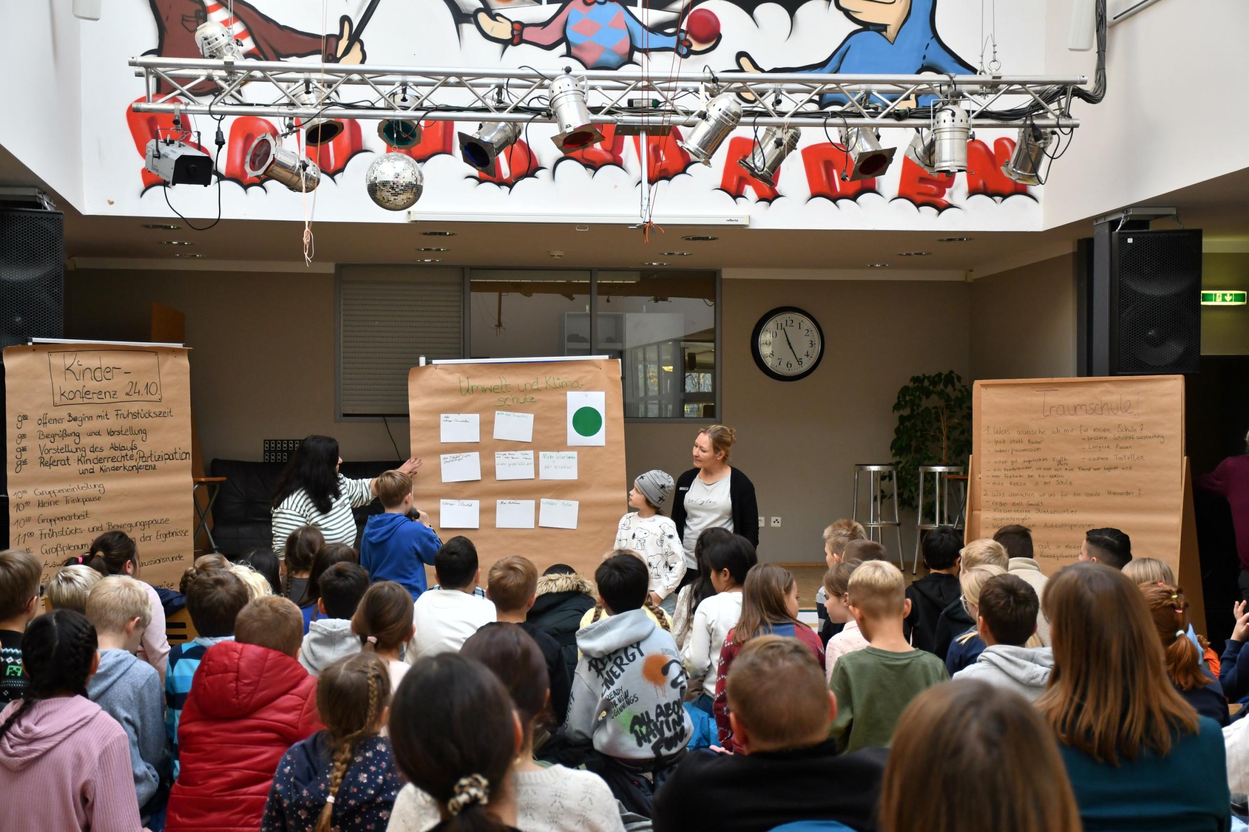 Ein Mädchen und ein Junge stehen vor einer mit einem breiten Faserstift beschriebenen Planwand und sprechen in ein Mikrofon.