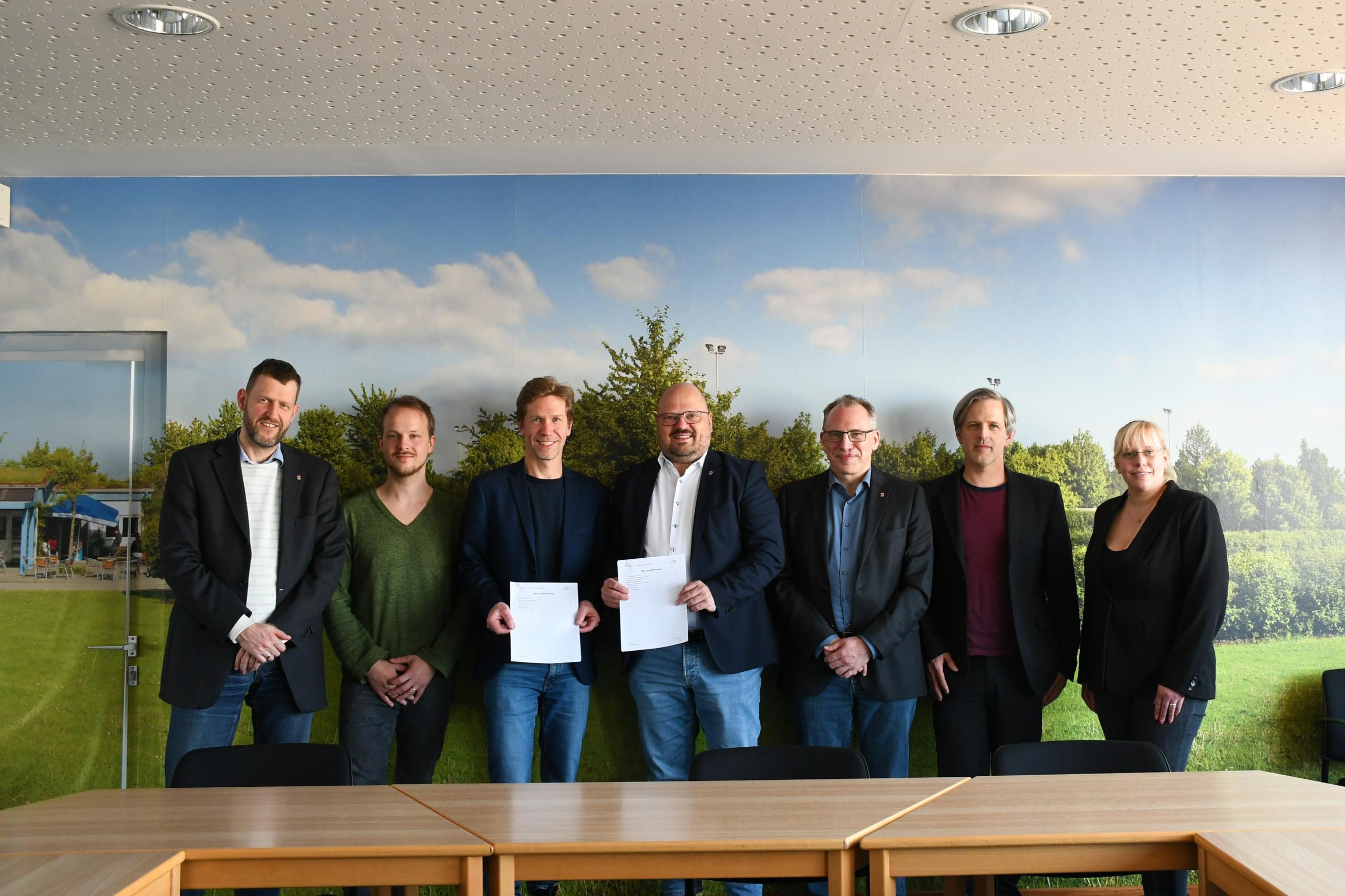 Eine Gruppe Menschen steht vor einer Fototapete mir einer Parklandschaft. Zwei von ihnen halten Verträge in den Händen.