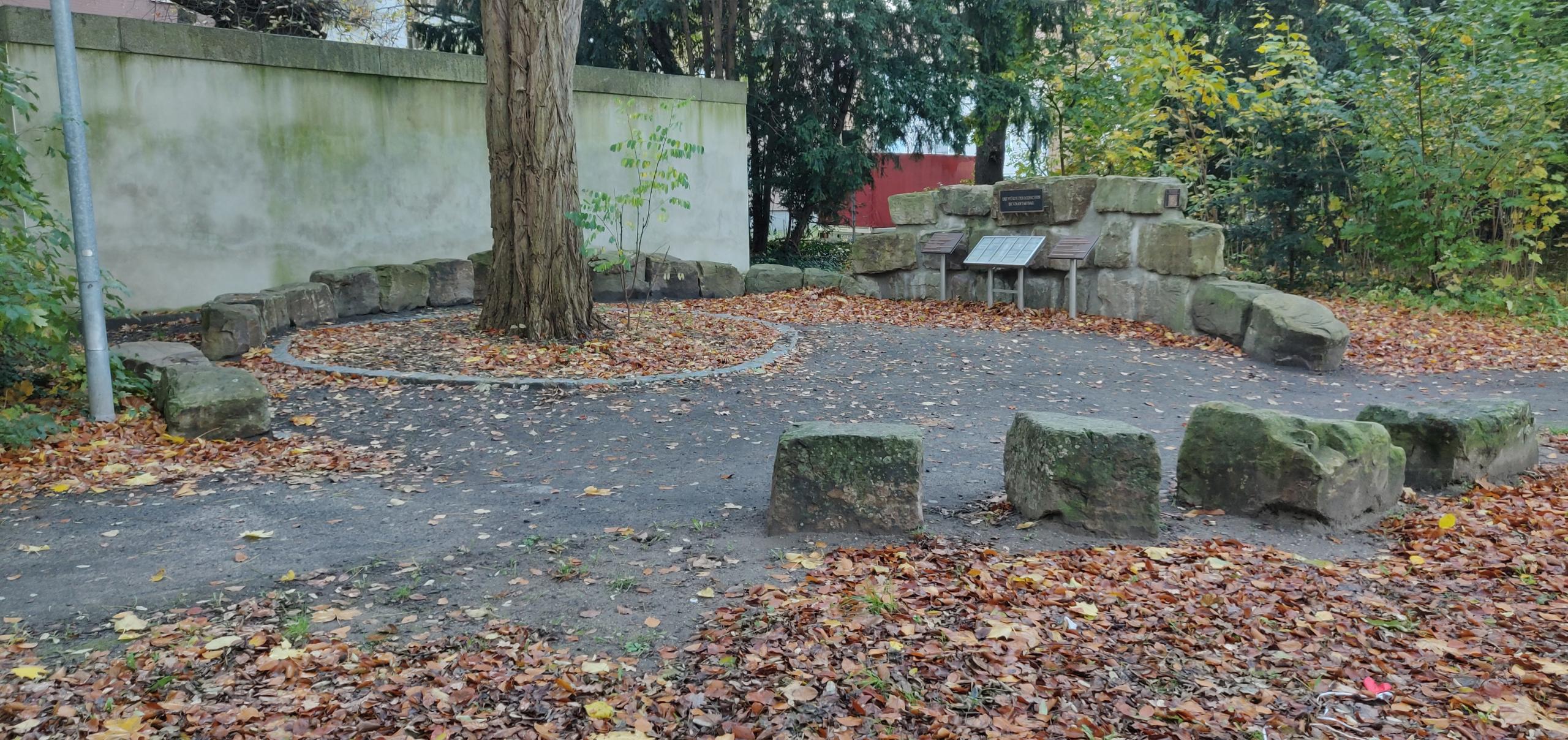 Kreis aus unbehauenen Natursteinen auf der Rückseite vom Ehrenmal in Alt-Laatzen. An einem Punkt des Kreise sind mehrere der unbehauenen Natursteine aufeinander gestapelt. An den Steinen ist zentral ein Bronzeschild angebracht. Davor steht zentral eine Metallkassette mit Toten Gedenksteine. Rechts und links von der Metallkassette steht je eine Bronzetafeln mit Inschrift.