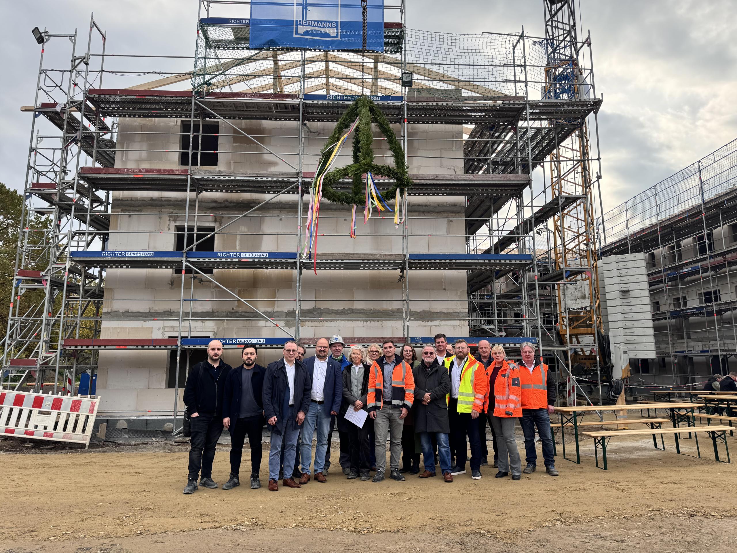 Eine größere Gruppe Menschen stehen vor einem mehrstöckigen Rohbau, über ihnen hängt der Richtkranz an einem Kran.