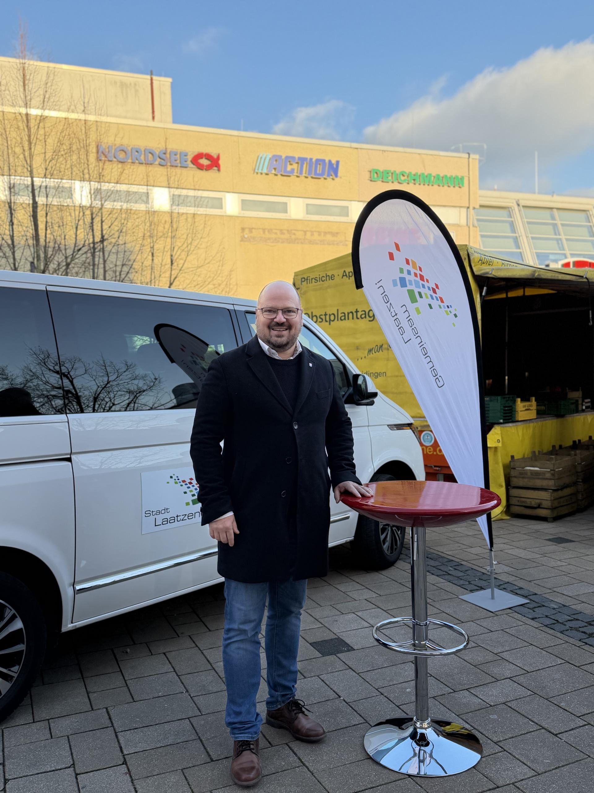 Ein Mann steht auf einem Platz neben einer Beachflag und einem Stehtisch.
