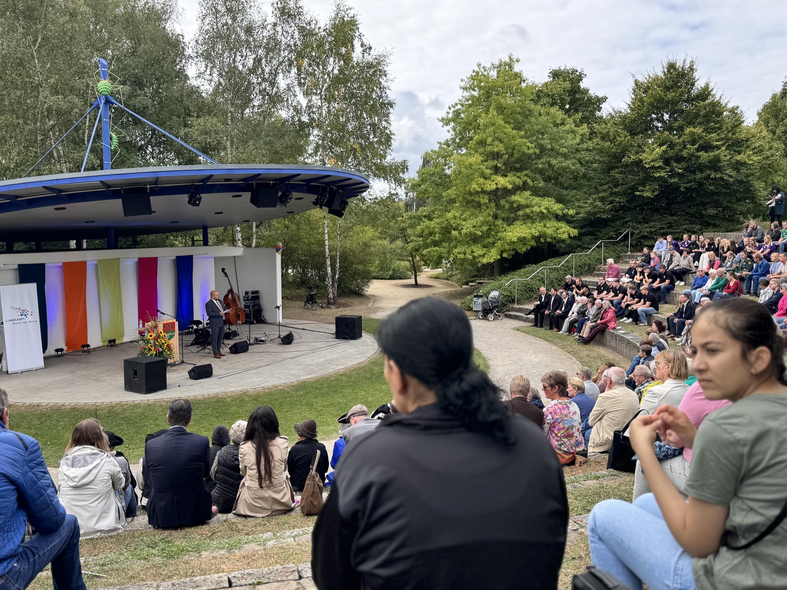Blick auf die Kulturbühne im Park der Sinne. Bürgermeister Kai Eggert spricht. Viele Personen sitzen im Publikum.