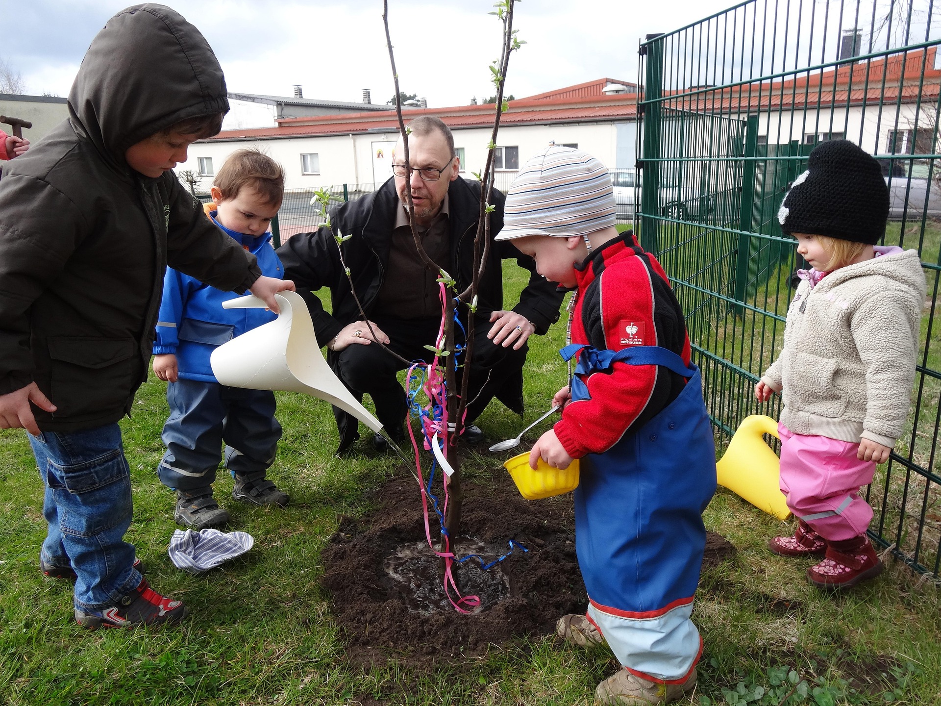 Vier kleine Kinder stehen mit ihrem Erzieher an einem frisch gepflanzten Baum und gießen diesen.