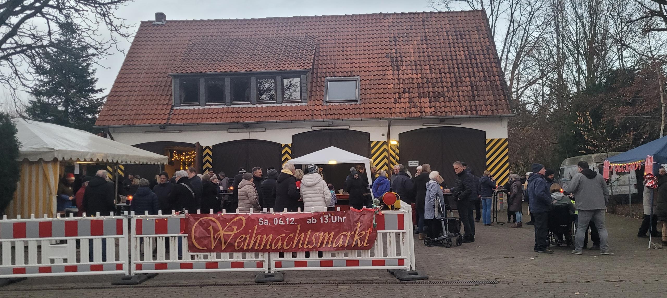 Blick auf das Gelände der alten Feuerwache Gleidingen. Im Vordergrund sind Menschen und Stände hinter einer Absperrung mit dem Banner "Weihnachtsmarkt" zu sehen. Im Hintergrund befindet sich das Gebäude der alten Feuerwache Gleidingen.