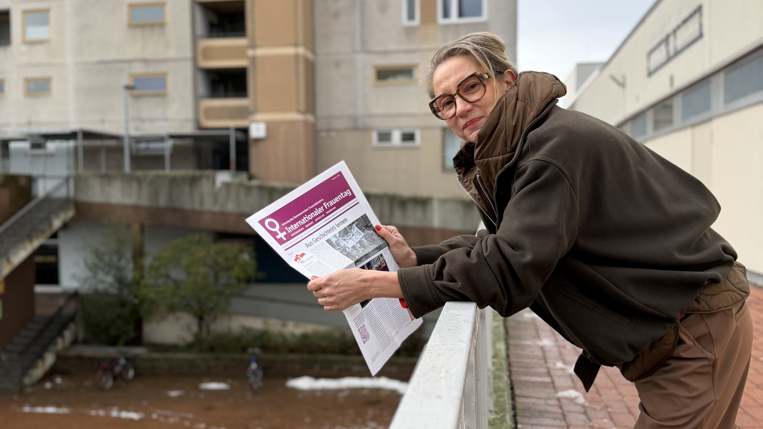 Eine Frau mit festem Blick lehnt über eine Brüstung, in der Hand hält sie eine Zeitung mit violettem Deckblatt.