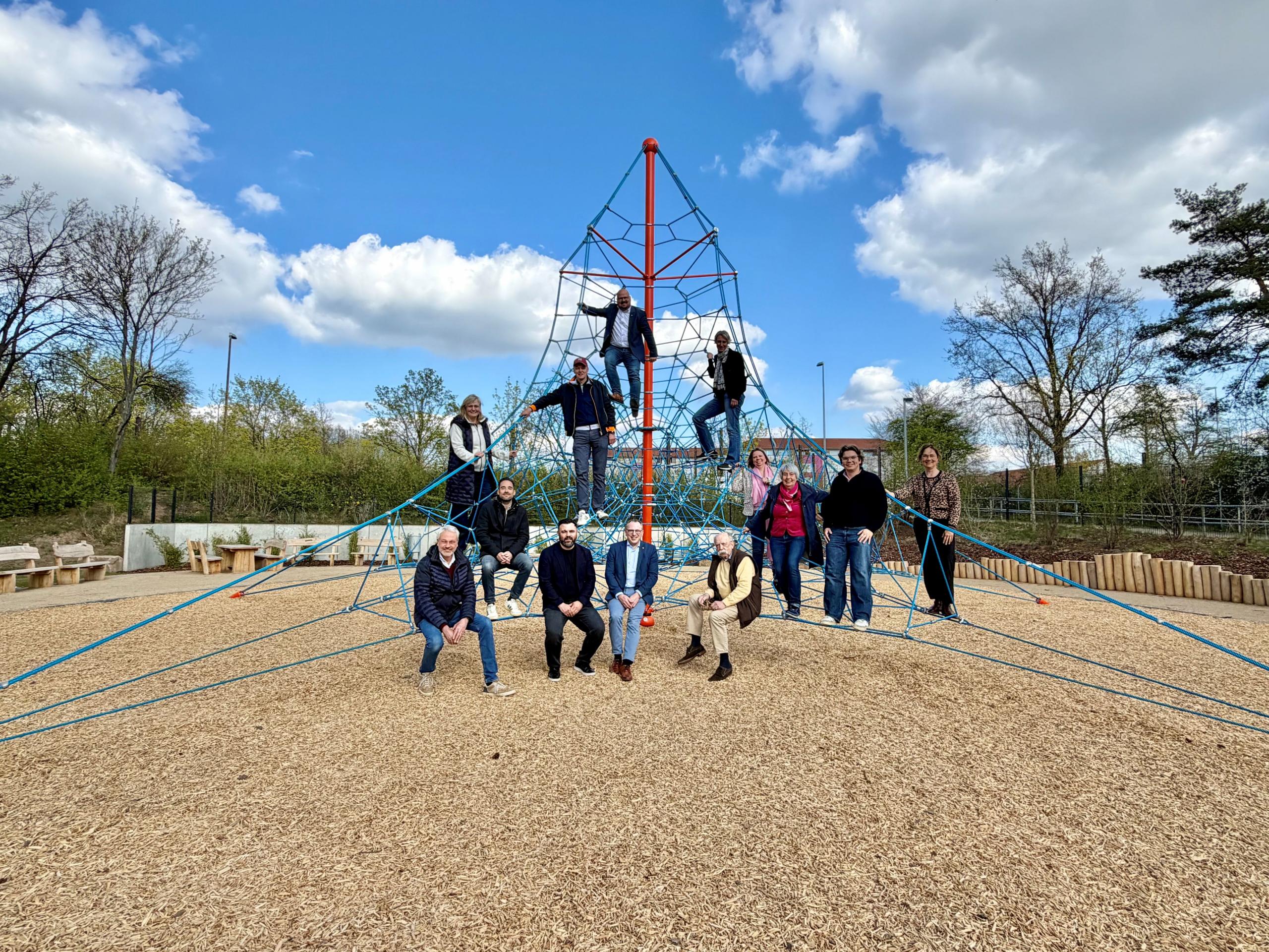 Herzstück des neuen Außenbereichs: die 7,2 Meter hohe Seilpyramide zum Klettern. Bürgermeister Kai Eggert und stellv. Direktorin Melanie Winter sowie Vertreterinnen und Vertreter von Stadtverwaltung, Schule, Politik und Bauunternehmen kamen zusammen, um das jüngst fertiggestellte Außengelände des Henry-Korman-Hauses zu besichtigen. ©Stadt Laatzen Eine Gruppe von 13 Personen hat eine Seilpyramide auf einem Spielplatz erklommen und sie schauen freundlich in die Kamera.