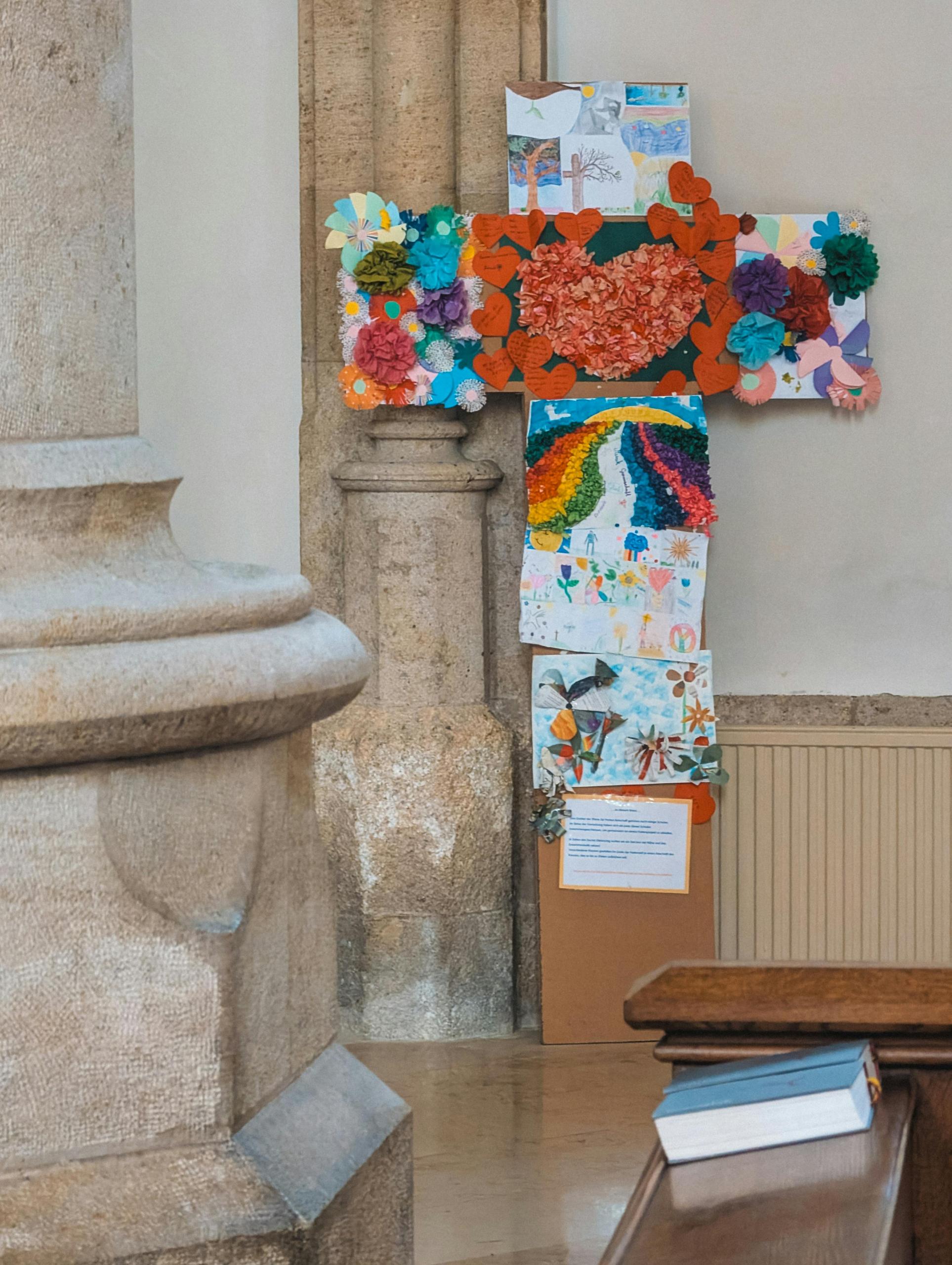 Sympolisches Kreuz aus bunt gemalten Kinderzeichnungen in einer Kirche. Eine Steinsäule auf der linken Seite, rechts eine Sitzbank mit Gesangsbuch.