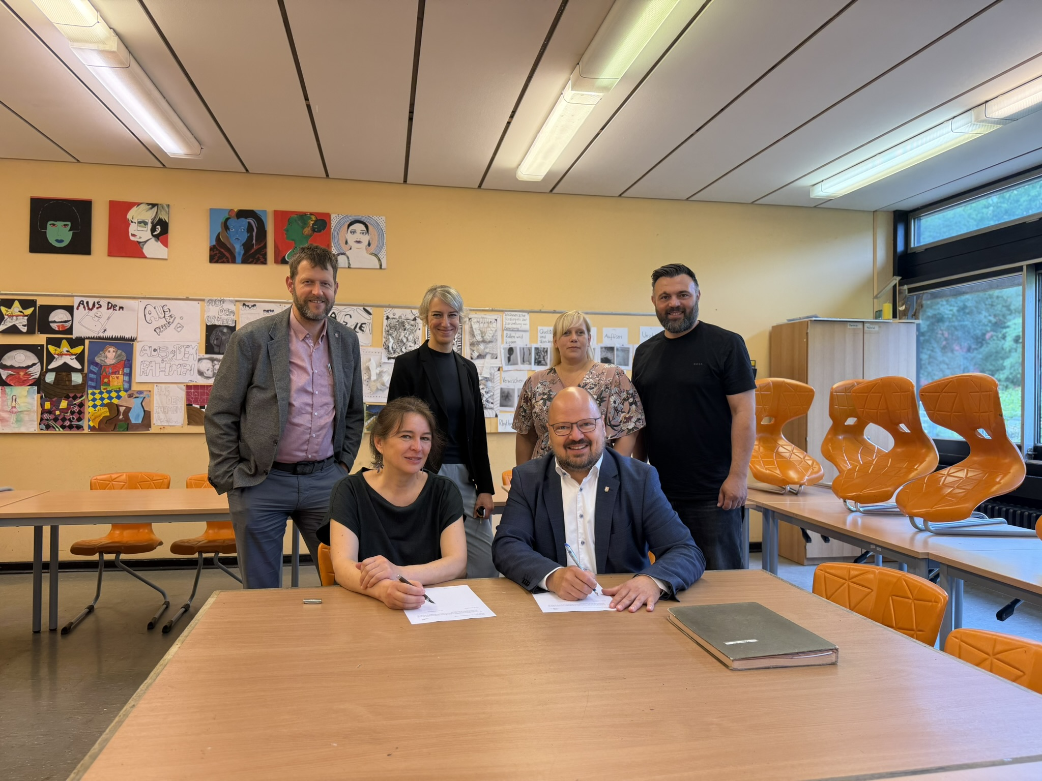 Startschuss für die Sanierung des C-Trakts der Albert-Einstein-Schule: Bürgermeister Kai Eggert und Kristina Gronau vom Planungsbüro Jensen Gronau Architekten unterzeichnen den Vertrag über die Planungsleistungen. Mit dabei sind Stadtrat Hauke Schröder (hinten v. l.), die stellvertretende Schulleiterin Melanie Winter sowie die Leiterinnen und Leiter der städtischen Fachbereiche Bauen, Jessica Kaußen, und Bildung, Ali Kara. ©Stadt Laatzen Bürgermeister Kai Eggert und Planerin Kristina Gronau unterzeichnen in einem Klassenraum den Vertrag zur Sanierung des C-Trakts der Albert-Einstein-Schule. Hinter ihnen stehen weitere Vertreterinnen und Vertreter von Schule und Stadtverwaltung.