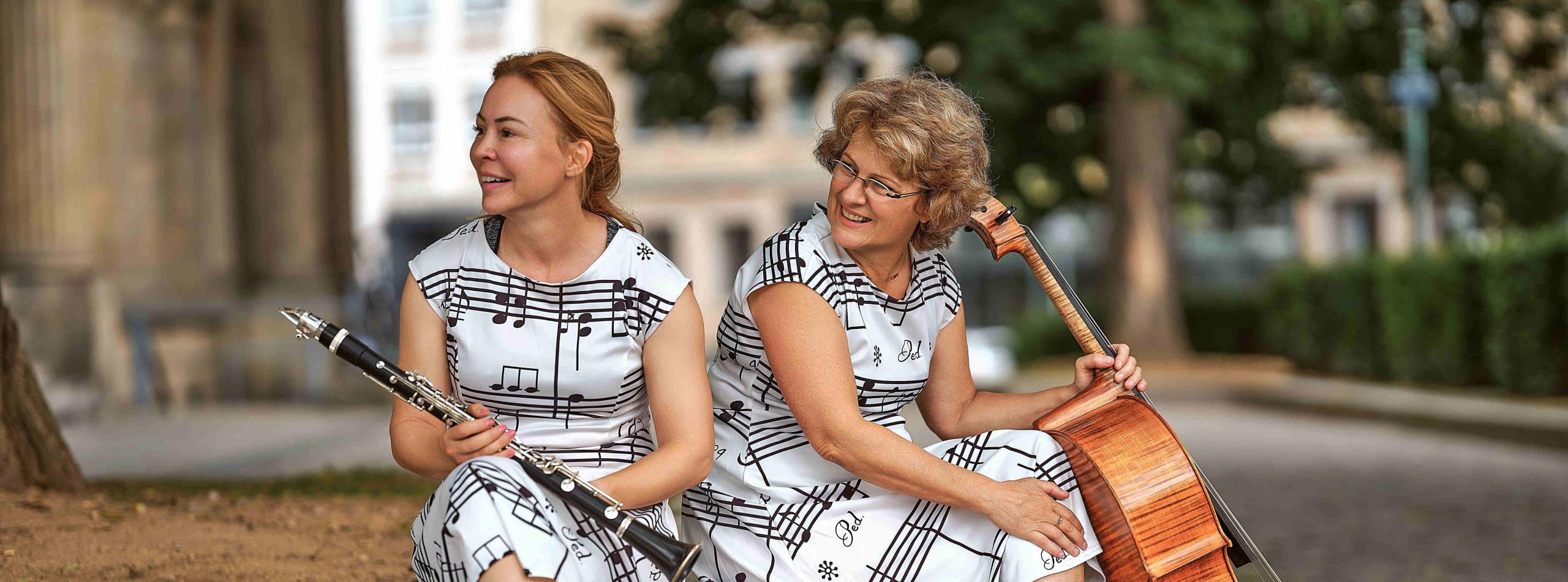 Zwei Frauen mit identischen Kleidern mit Musiknoten sitzen auf einem urbanen Pflanzbeet aus Beton. Die eine hält ihre Klarinette in der Hand, die andere ihr Cello.
