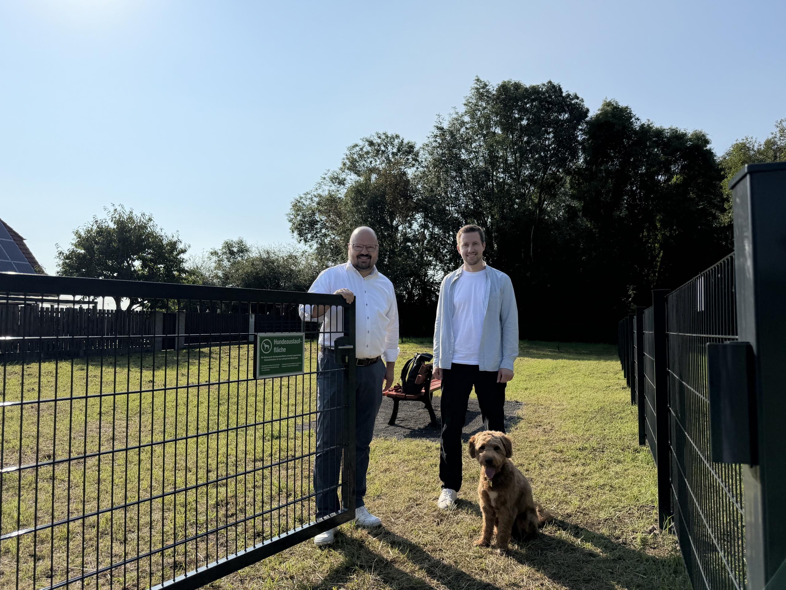 Platz zum Spielen und Toben: Bürgermeister Kai Eggert (l.) und Marvin Meyer, Leiter des städtischen Grünflächen-Teams, geben die über 500 Quadratmeter große Hundeauslauffläche am Sirupweg in Rethen offiziell frei. Hündin Maily wartet auf ihren Einsatz. ©Stadt Laatzen Bürgermeister Kai Eggert und Leiter des städtischen Teams Grünflächen stehen an einem geöffneten Tor vor einer Hundewiese. Davor sitzt ein mittelgroßer Hund mit braunem Fell..