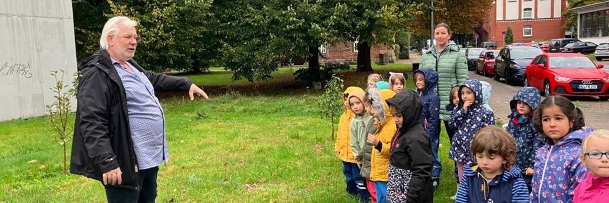 Eine Gruppe von kleinen Kinder stehen in einer Gruppe. Vor ihnen ein Mann, der auf einer Wiese steht und den Kinder etwas erzählt.