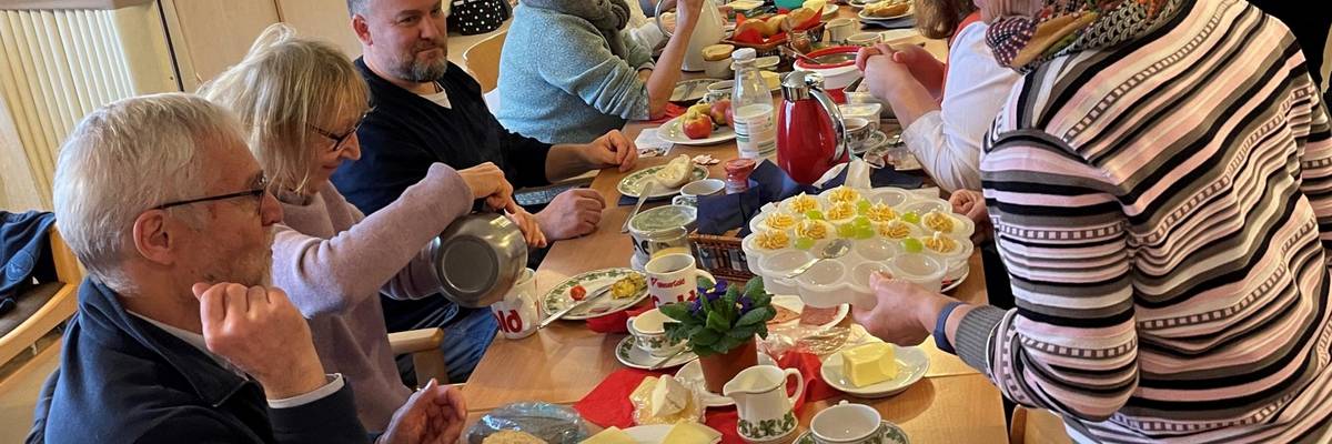 Eine Gruppe von Menschen sitzt an einem langen Tisch und frühstückt. Der Tisch ist gedeckt mit Obst, Brötchen und weiteren Lebensmitteln.