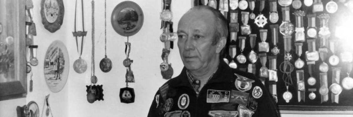 Herr mit zahlreichen Abzeichen an seiner Jacke und im Hintergrund an der Wand.