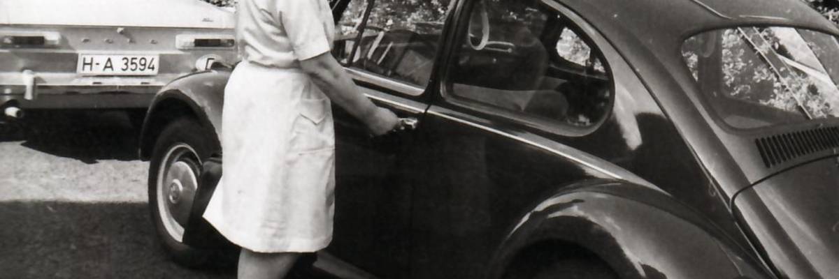 Krankenschwester öffnet Tür von Auto, davor ein anderes Auto, im Hintergrund ein Gebäude