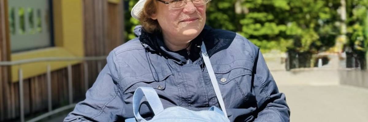 Bild Instagram-Beitrag © Stadt Laatzen Eine Frau sitzt in einem Rollstuhl im Freien. Sie trägt einen hellen Sonnenhut, eine Brille, eine dunkelblaue Jacke und eine hellblaue Tasche. Sie blickt leicht zur Seite und lächelt.