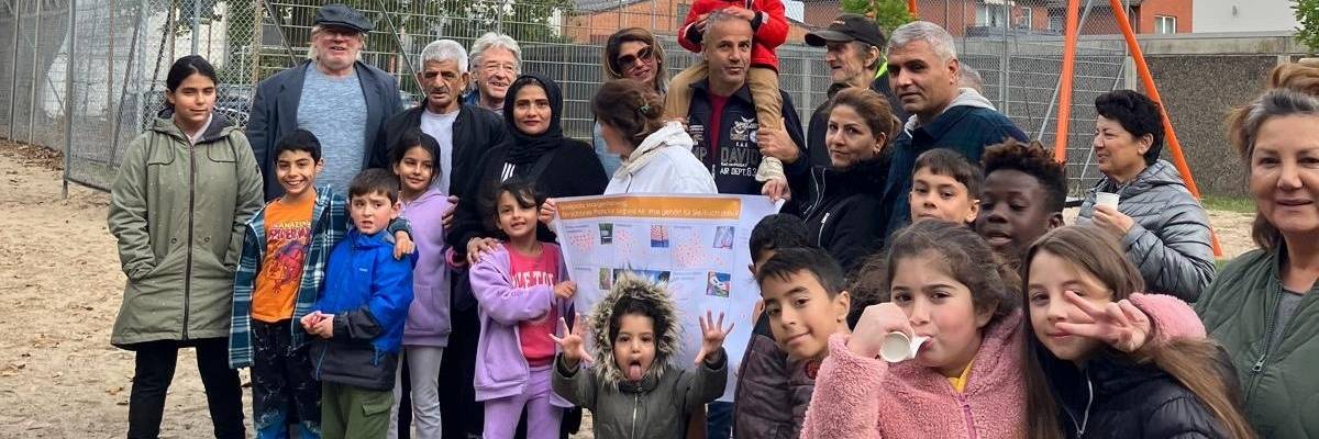 Eine Gruppe Frauen, Männer und Kinder auf einem Spielplatz. Sie szehen vor einem Zaun.