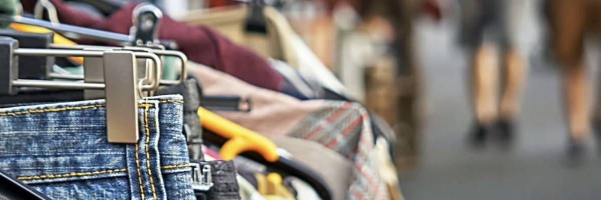 Bild Instagram-Beitrag © Canva Flohmarkt-Szene mit Kleiderstange im Vordergrund, an der verschiedene Kleidungsstücke hängen. Im Hintergrund sind Personen sowie weitere Verkaufsstände sichtbar.