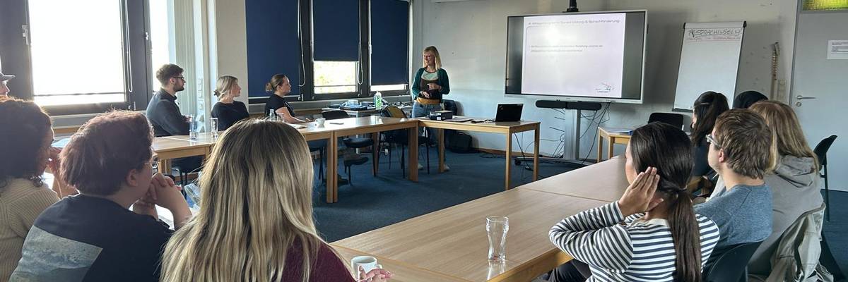 Die Referentin Maike Waßmann steht am Whitboad und präsentiert eine Powerpoint. Die Fachkräfte sitzten in U- Form zur Präsentation.