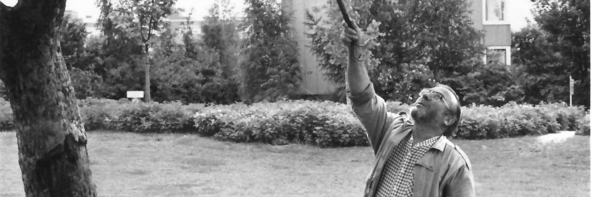 Mann zeigt mit Stab Richtung Baum, dahinter Schubkarre und Gießkanne auf einer Grünfläche, im Hintergrund ein Haus