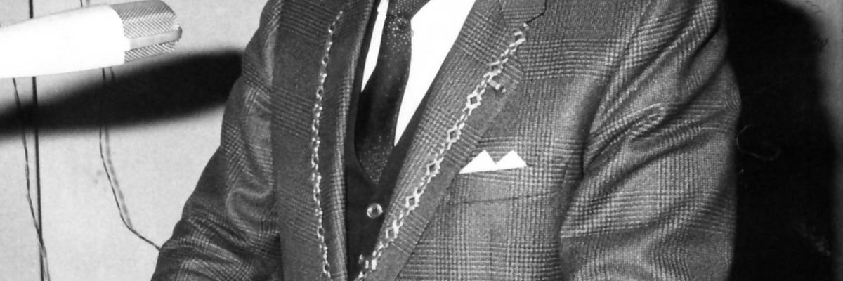 Mann mit Kette am Mikrofon, im Hintergrund ein Bild an der Wand