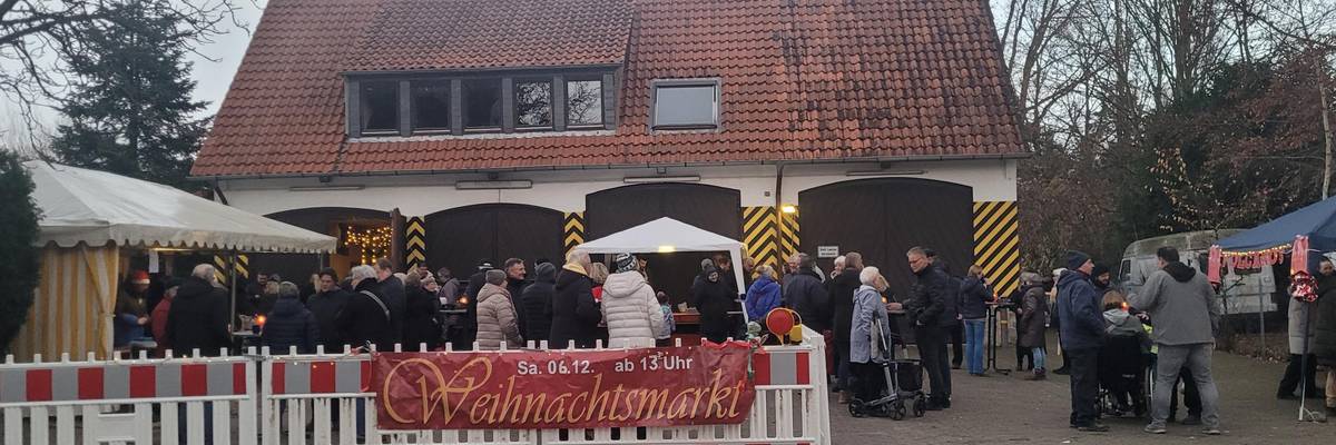 Blick auf das Gelände der alten Feuerwache Gleidingen. Im Vordergrund sind Menschen und Stände hinter einer Absperrung mit dem Banner "Weihnachtsmarkt" zu sehen. Im Hintergrund befindet sich das Gebäude der alten Feuerwache Gleidingen.