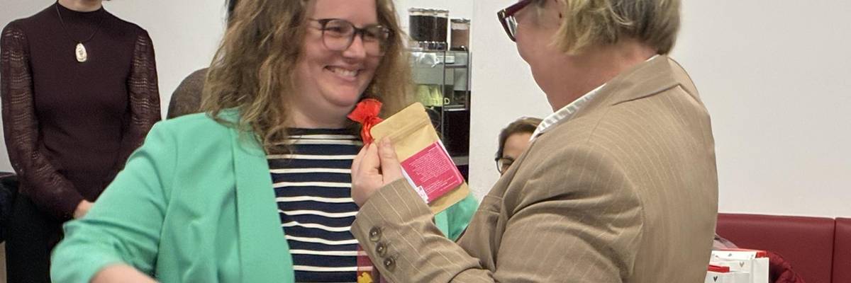 zwei Frauen stehen sich gegebüber. Die eine Frau überreicht das Zertifikat, ein Dokument, an die andere Frau.