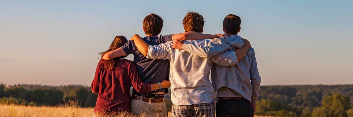 Vier Jugendlichen von hinten fotografiert, sitzen arm in arm auf einem Feld