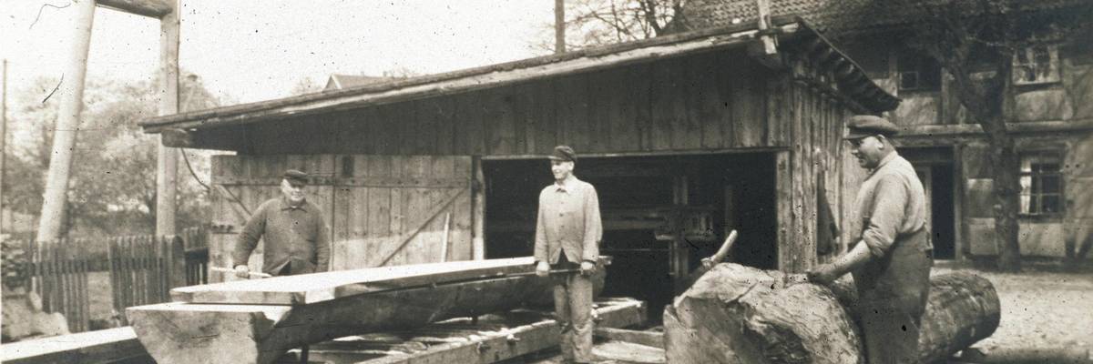Drei Männer arbeiten mit Holz. Vor einem der Männer liegt ein großer Baumstamm, zwei anderer stehen neben einem gesägten Brett.