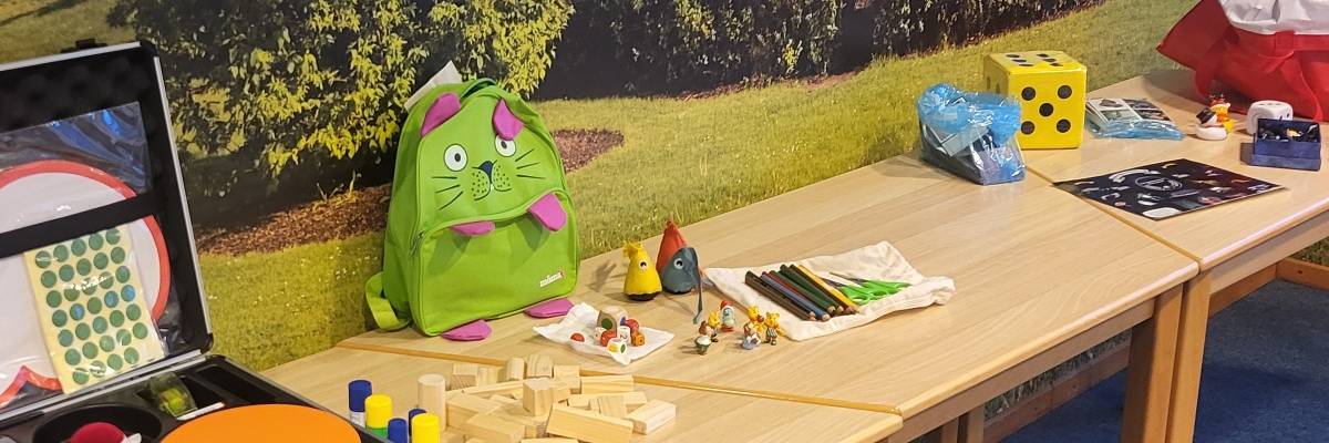 Auf drei Tischen stehen Spielmaterialien. Würfel, Spielfiguren, Aufkleber,...