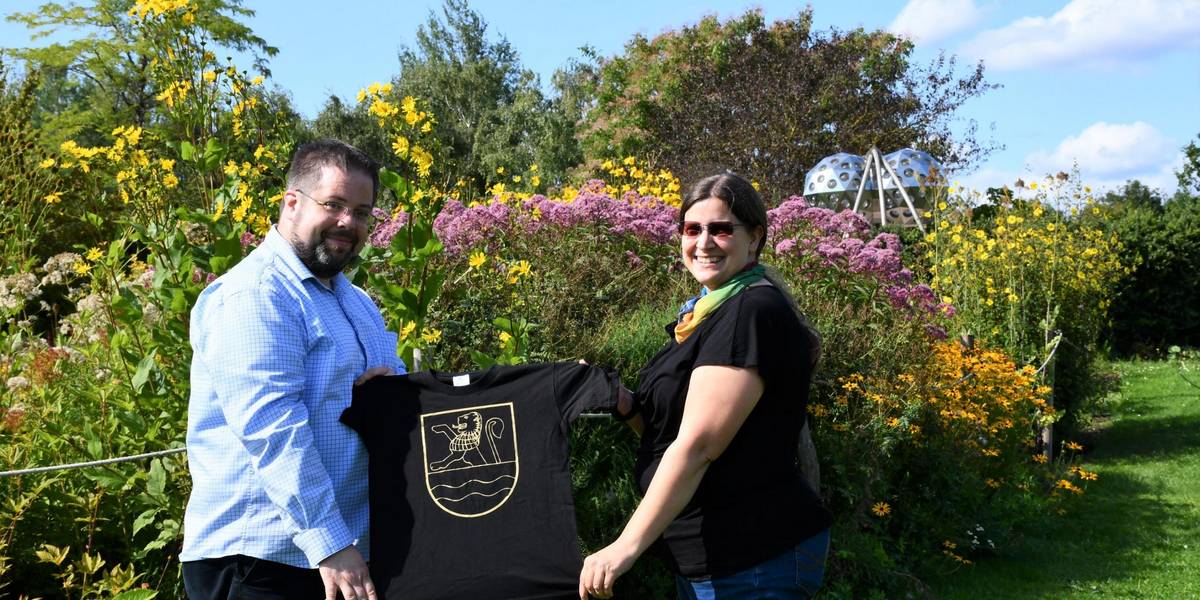 Zwei Personen, ein Mann und eine Frau stehen in einem Park. Hinter ihnen befindet sich ein buntes Blumenbeet. Sie halten ein schwarzes T-Shirt in die Höhe.