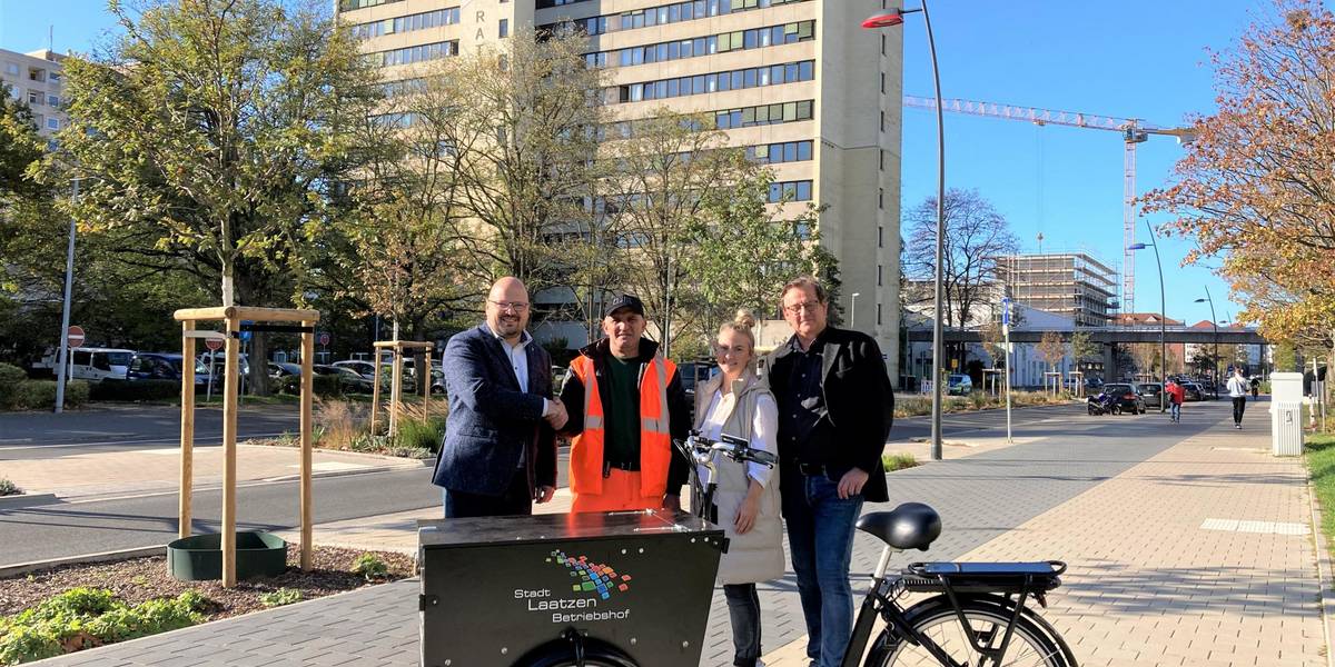 Bürgermeister Kai Eggert begrüßt, zusammen mit Juliane Erstling aus dem Team Tiefbau und Thomas Güllner vom Jobcenter, Laatzens ersten Stadtbildpfleger Ali Yildirim. © Stadt Laatzen/Anke Wesbrich Vier Menschen vor einem Lastenrad. Im Hintergrund ist eine Straße mit Brücke zu sehen.