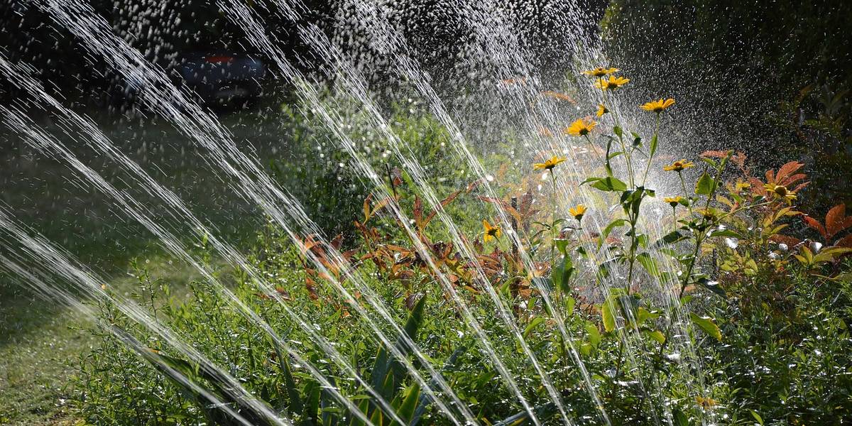 Gartensprenger steht an einem Blumenbeet mit gelben Blüten. Im Hintergrund ist eine Rasenfläche zu sehen.