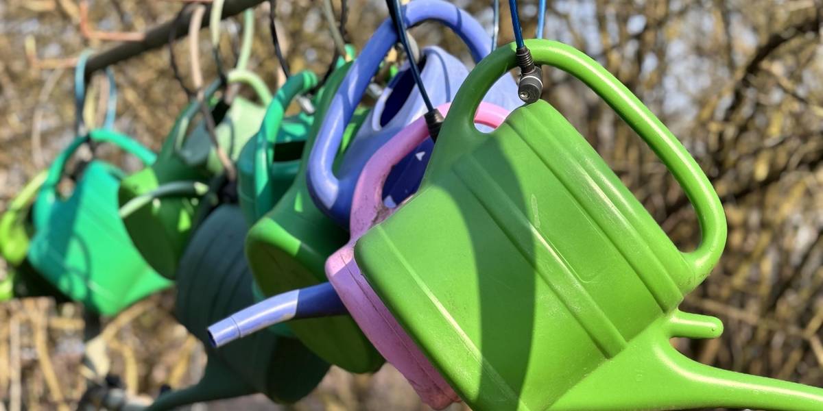 Eine Reihe bunter Gießkannen hängt nebeneinander. Alle Kannen sind mit Schössern angebunden.